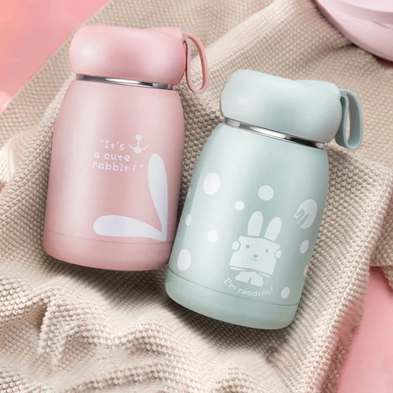 Two insulated cups, one pink and one light blue, with cartoon designs on a textured fabric background.